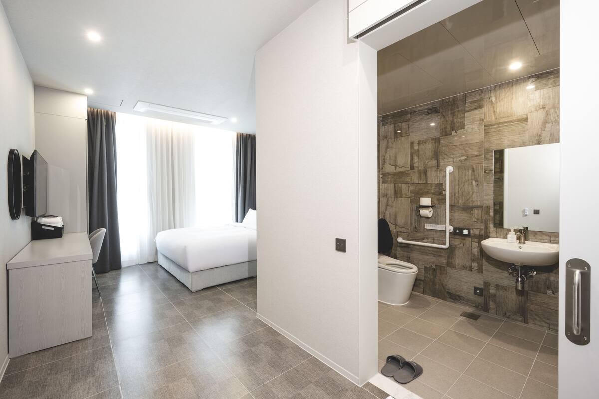 Quarto de hotel moderno com cama, escrivaninha, banheiro com chuveiro e vaso sanitário.
