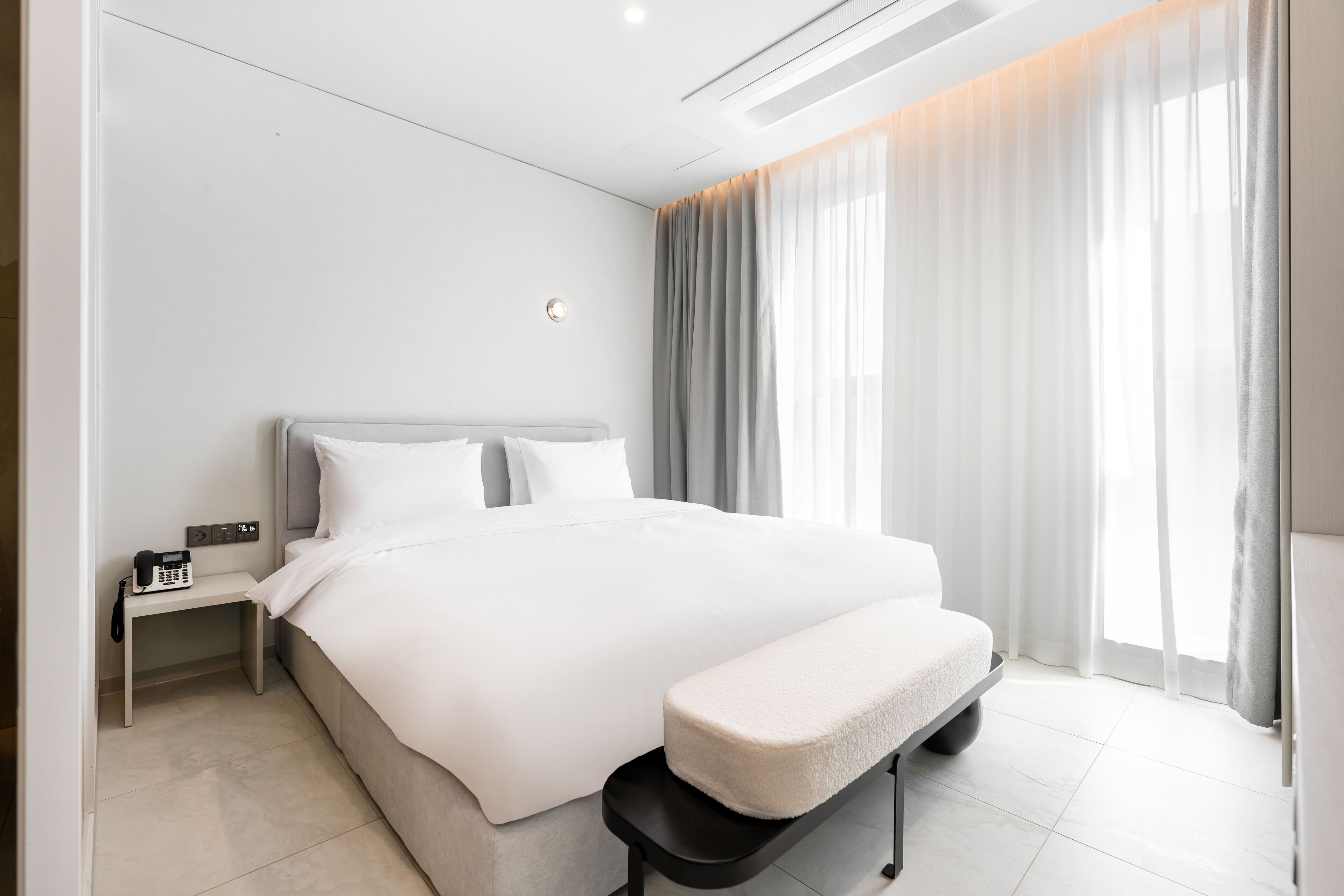 A modern hotel room with a large bed, bedside table, and a bench.