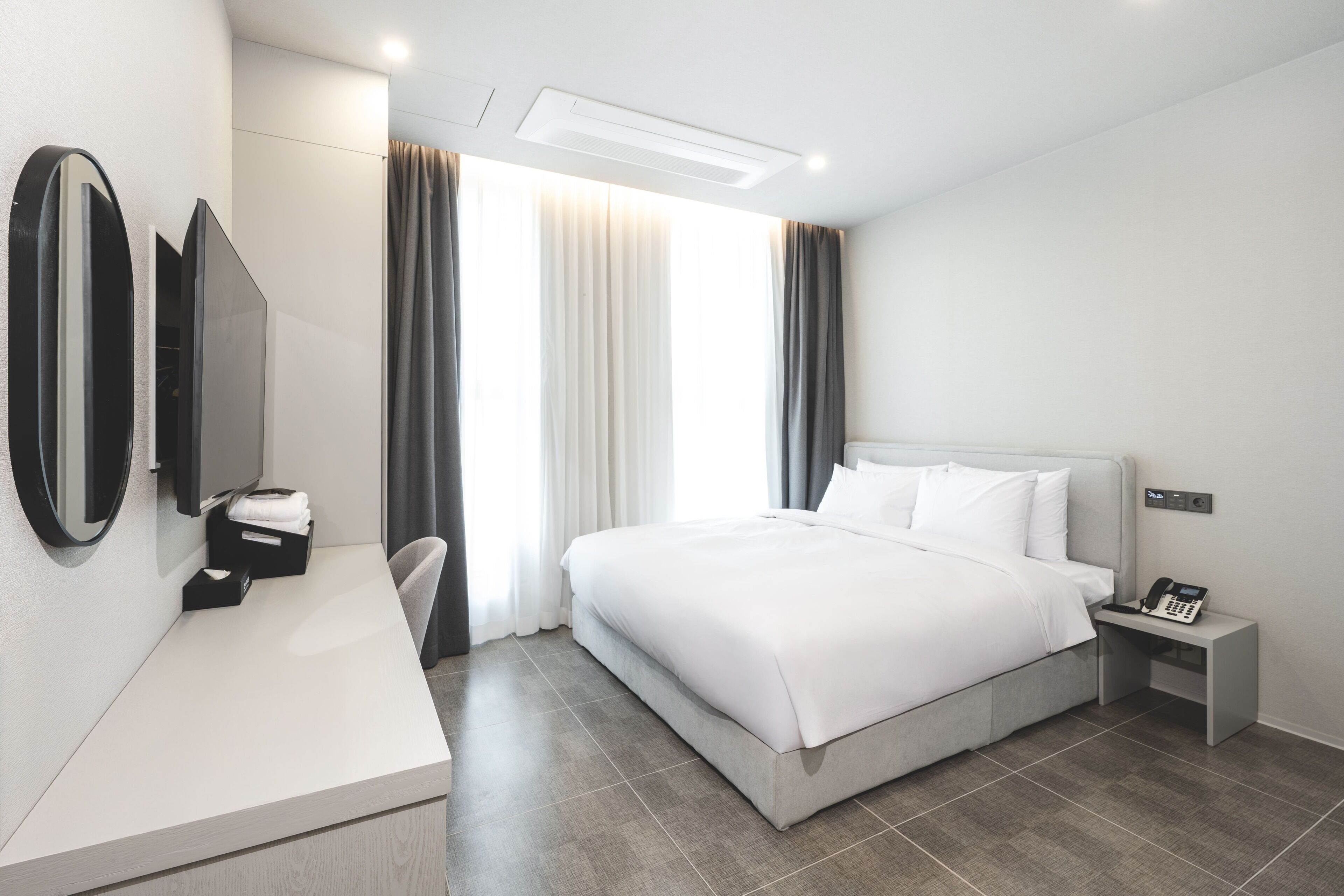 A modern hotel room with a large bed, a bedside table with a phone, a desk with a mirror, and a chair.