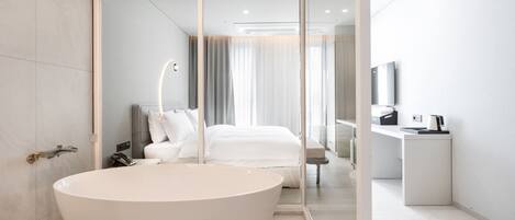 A modern bathroom with a freestanding bathtub, a shower area, and a visible bed in the background.