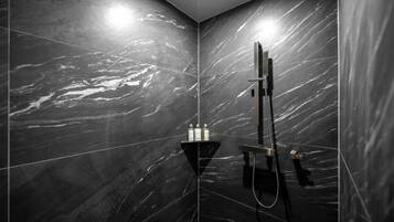 Un baño moderno con baldosas de mármol oscuro, ducha con cabezal de ducha manual y un estante pequeño con artículos de aseo.