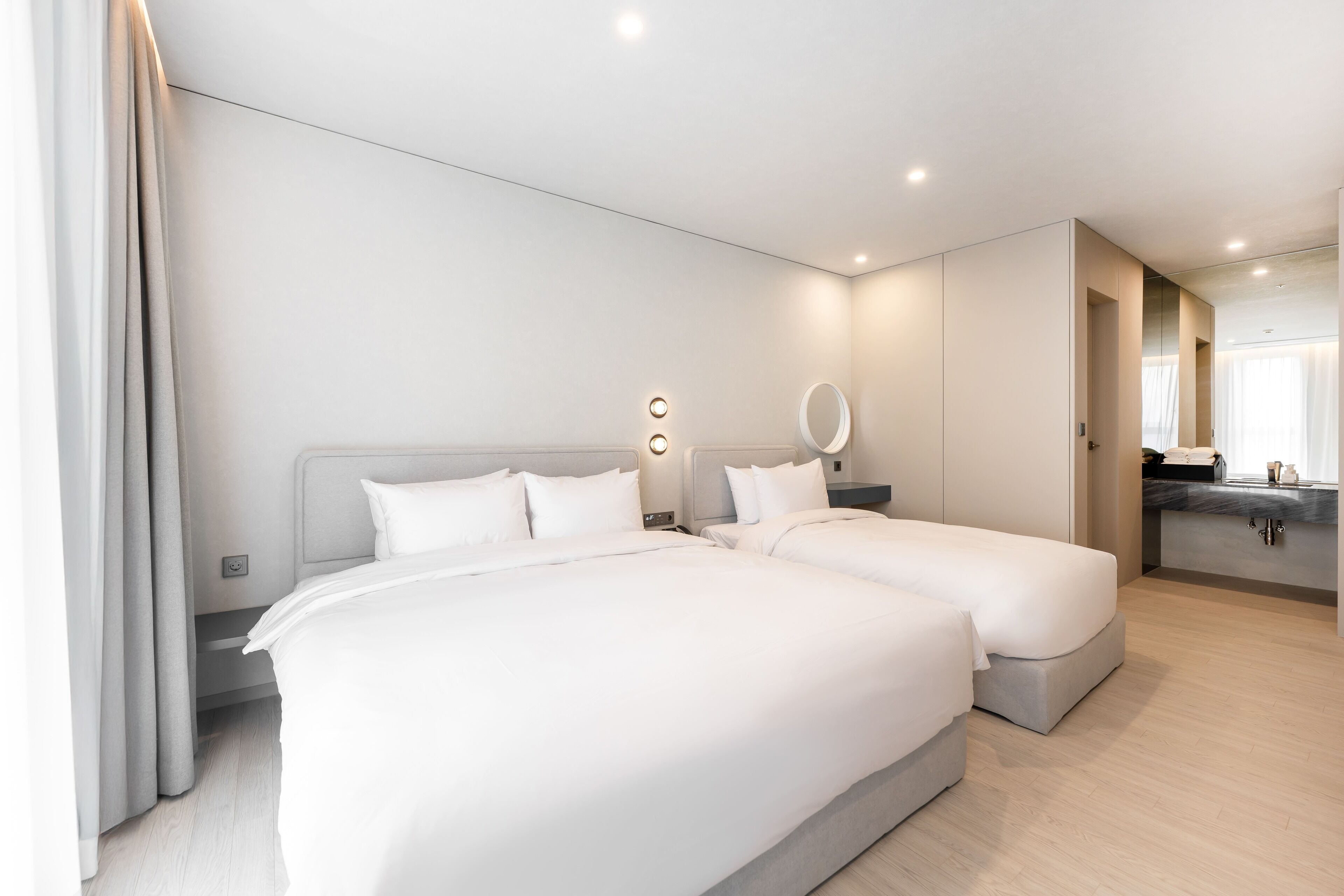 A modern hotel room with two beds, a built-in desk, and a bathroom with a mirror.