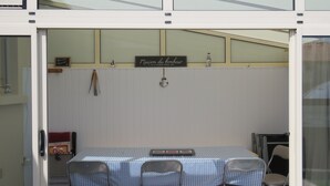 Une petite pièce fermée, avec une table et des chaises, une enseigne indiquant «Maison du Bonheur» et une petite étagère contenant divers objets.