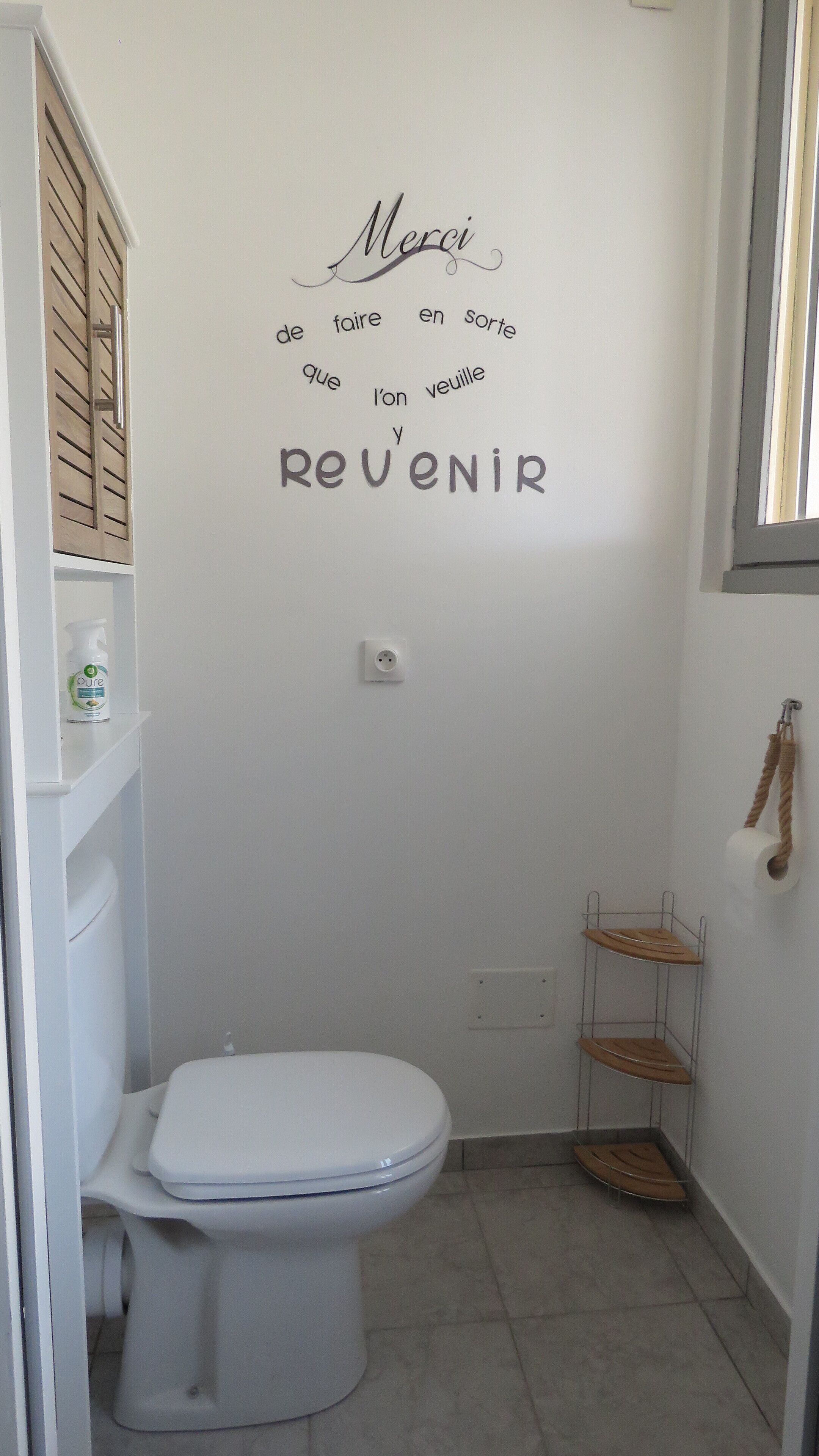 Een kleine badkamer met een wit toilet, een plankje met een fles, en een muur met een Franse tekst.