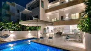 A modern outdoor pool area with lounge chairs and umbrellas, surrounded by greenery and a contemporary building.