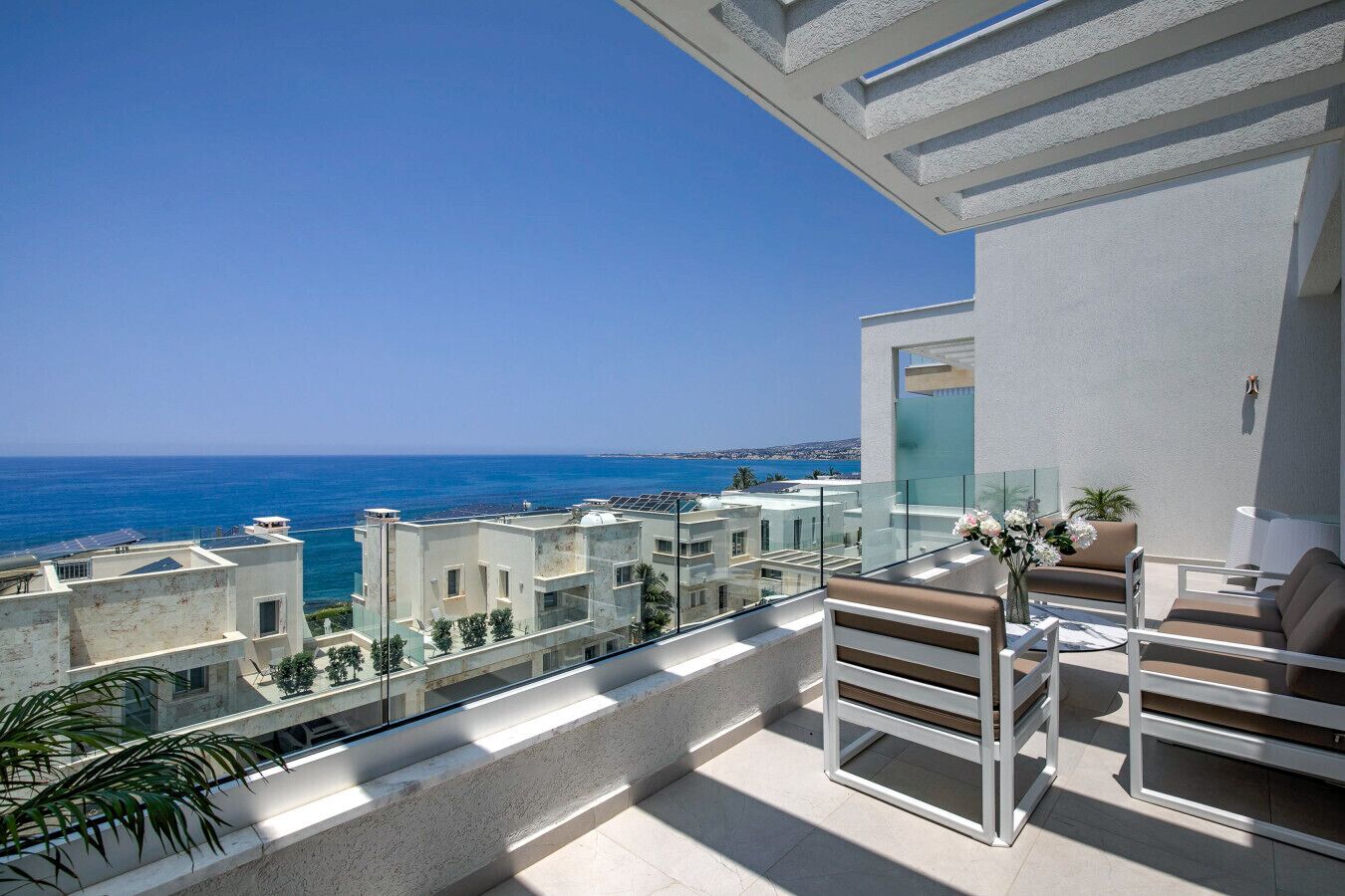 Un balcón con vistas al mar, barandilla de cristal y zona de estar con mesa y sillas.