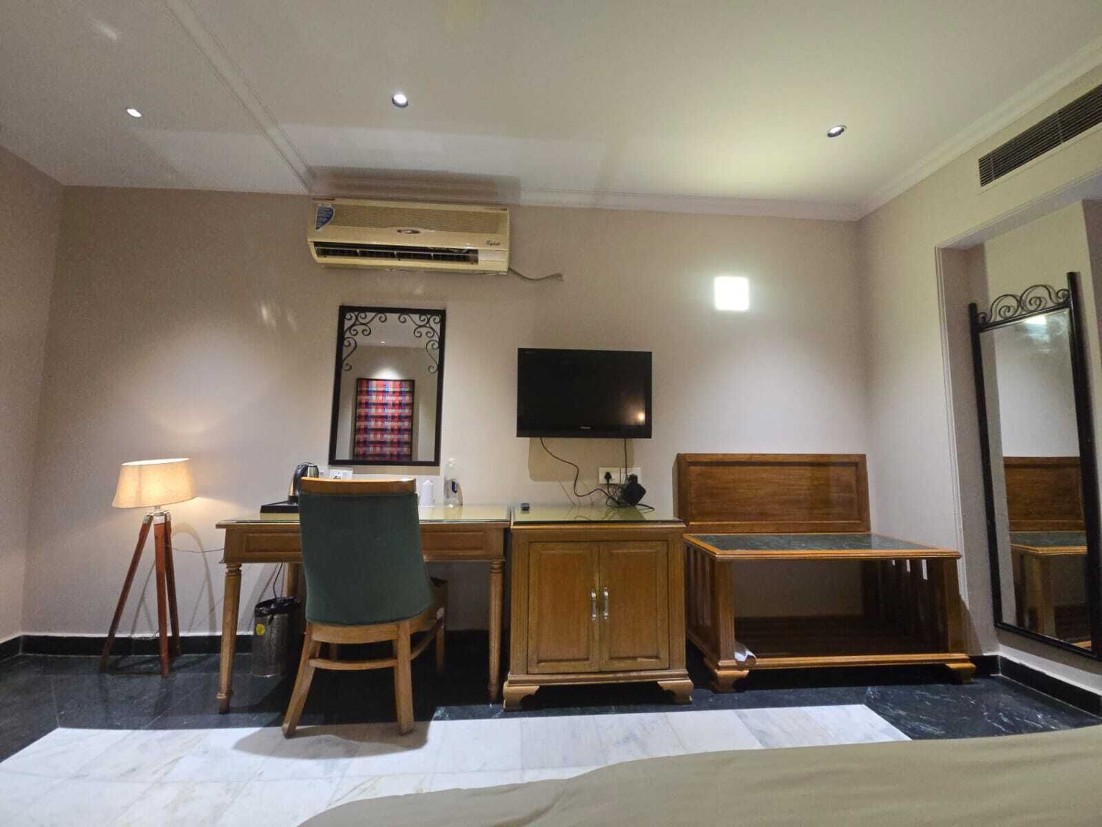 A hotel room with a bed, a desk with a chair, a television, an air conditioning unit, and a mirror.
