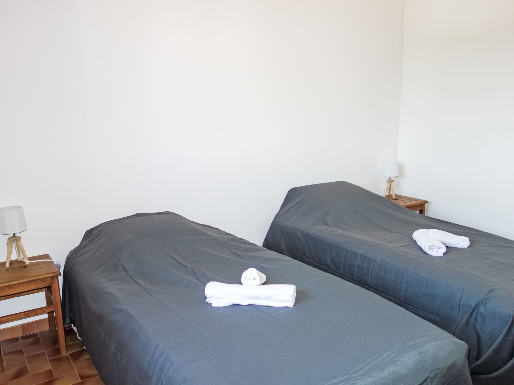 Two single beds with grey bedding and white towels folded on top. There is a wooden nightstand with a lamp on the left side.