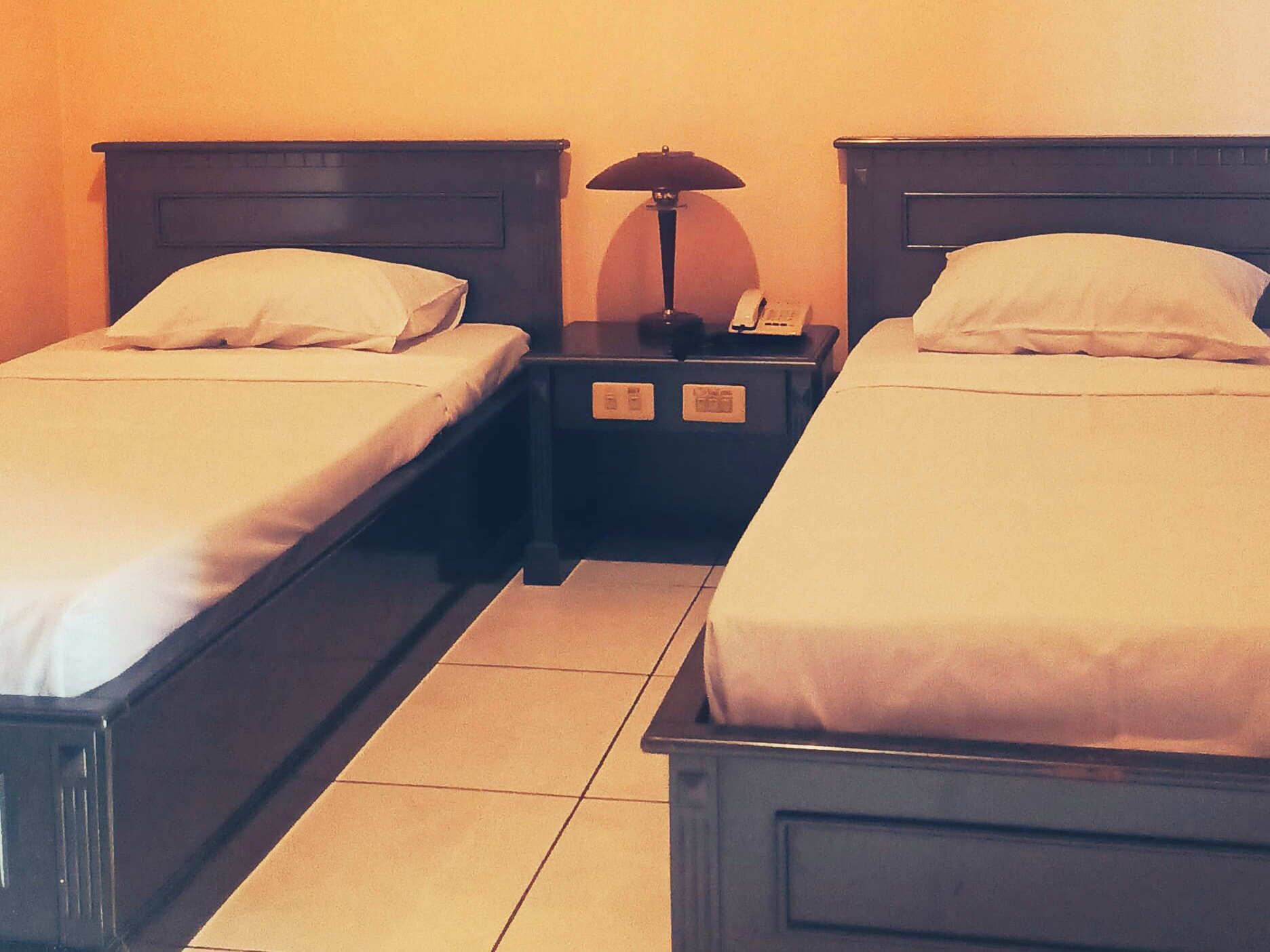 A hotel room with two single beds, each with a pillow and a nightstand with a lamp.