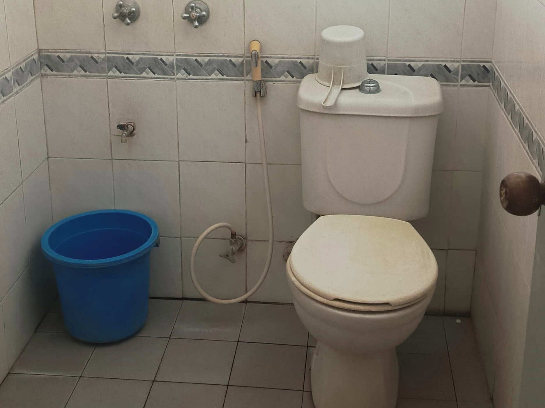 Um banheiro pequeno com vaso sanitário, um balde azul e suporte para papel higiênico.