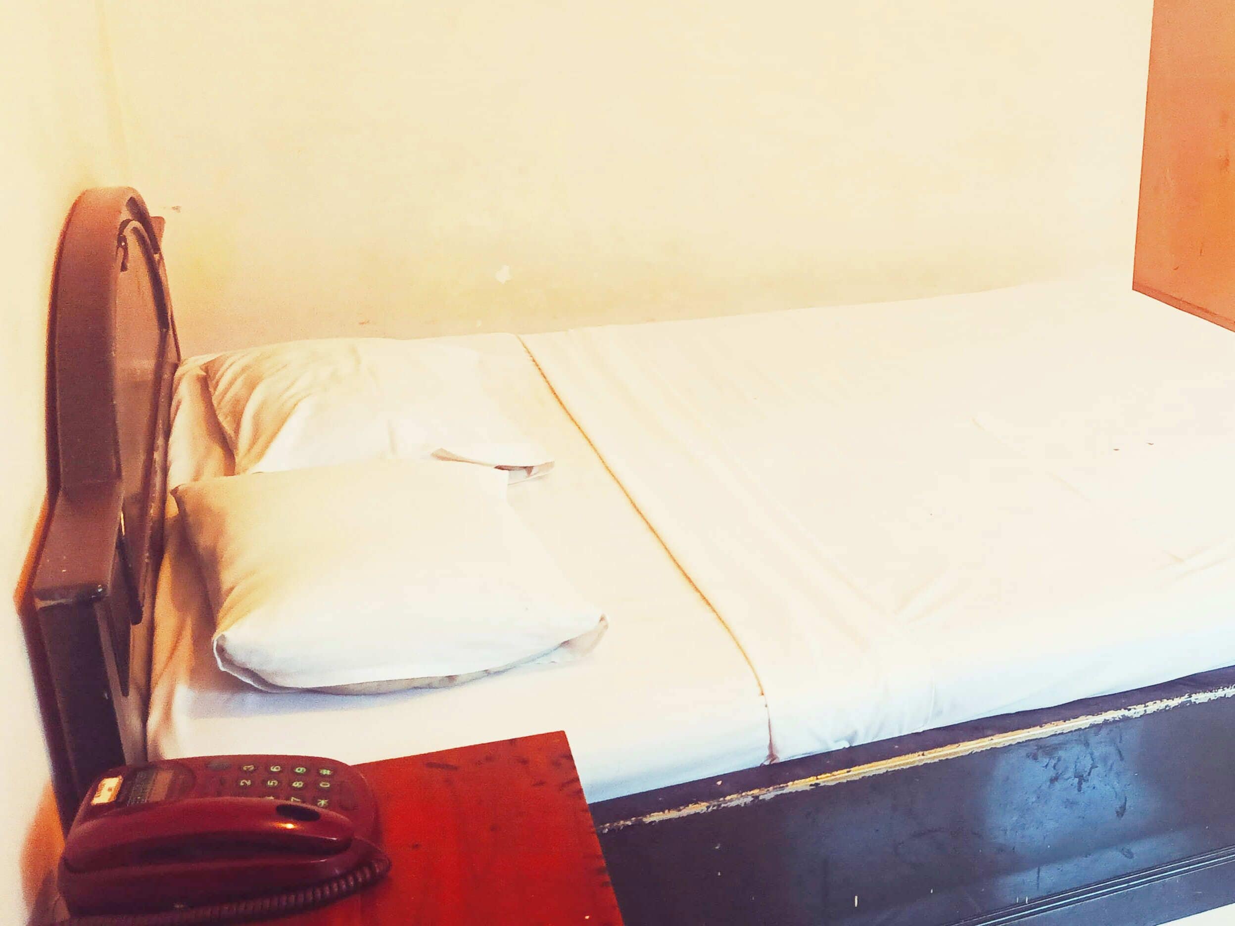 A small, simple room with a bed, a pillow, a red object, and a telephone on the floor.