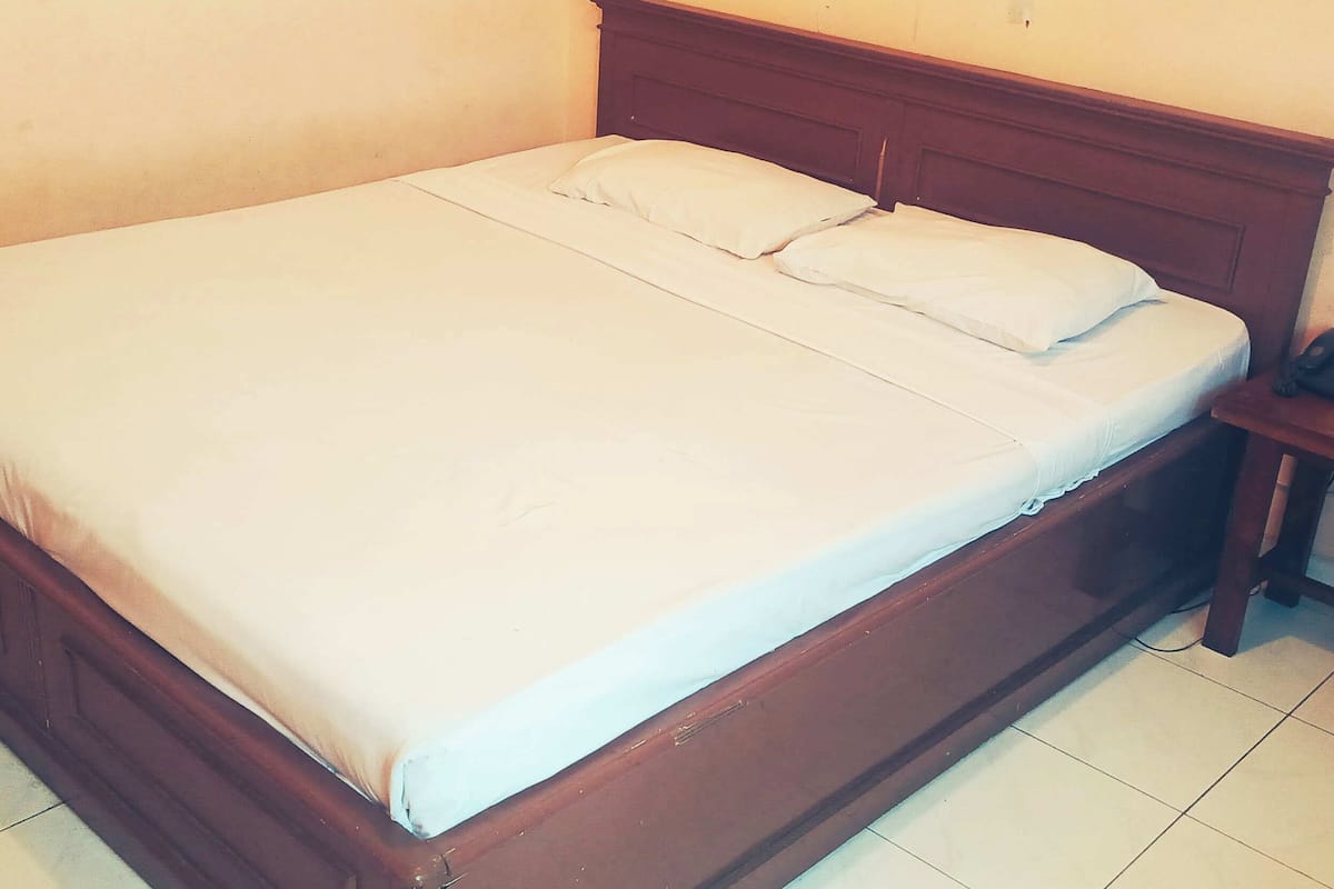 A single bed with a wooden headboard and white bedding in a room with plain walls and tiled flooring.