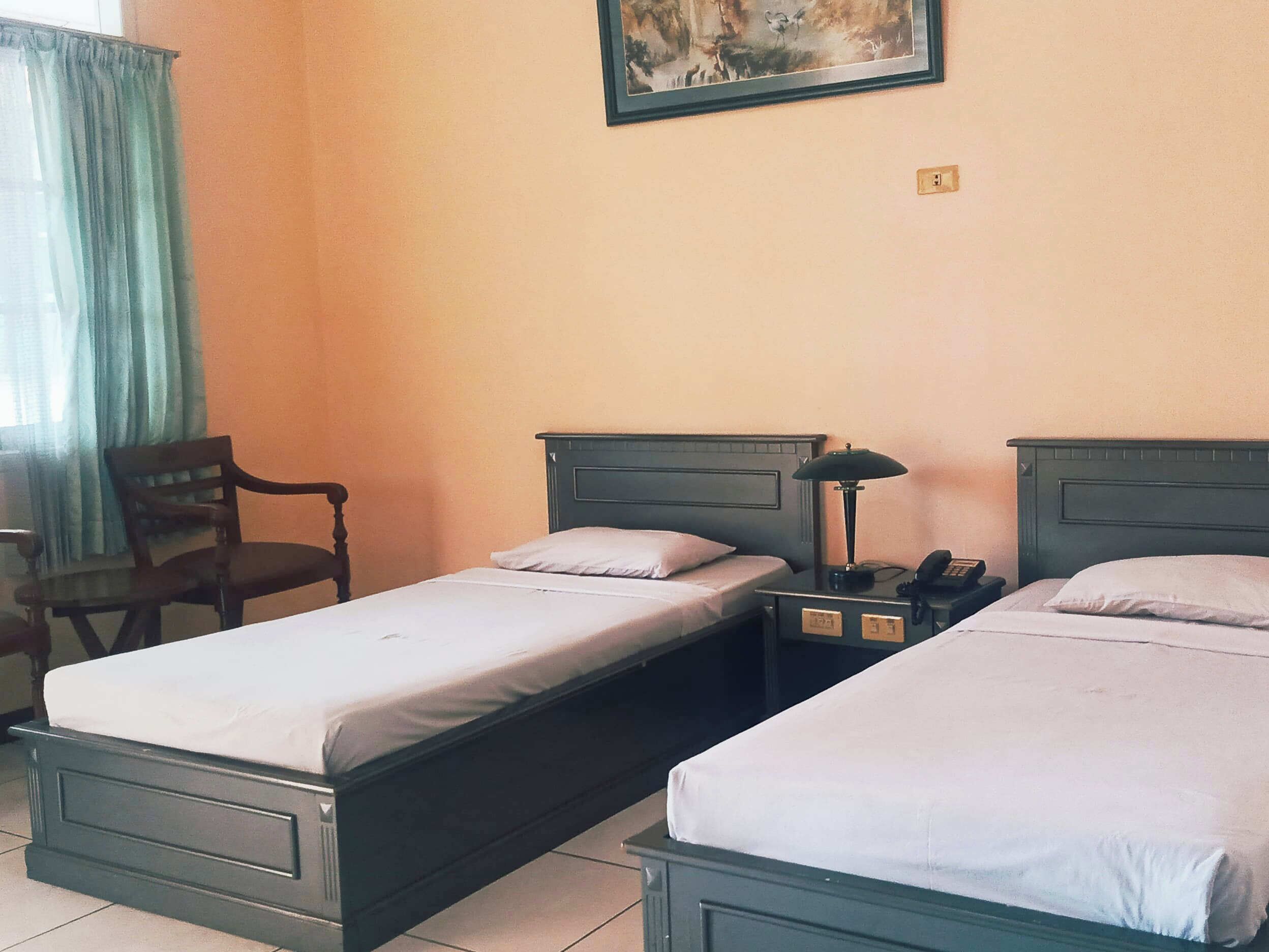 A hotel room with two single beds, a nightstand with a lamp, a chair, and a painting on the wall.
