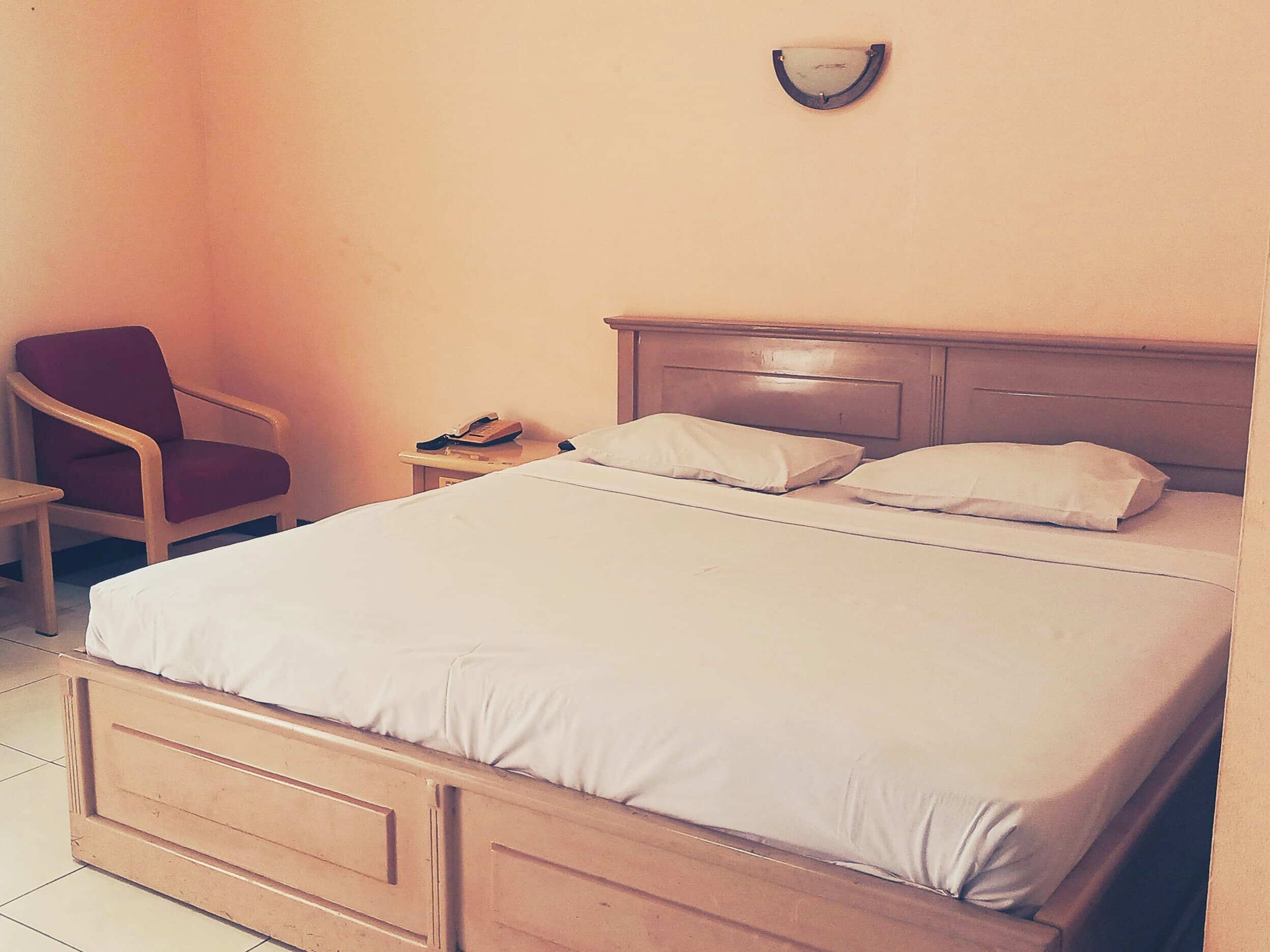A neatly made bed with a wooden headboard, a red chair, and a bedside table with a phone.