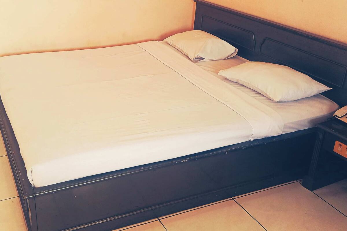 A single bed with white linens and two pillows, a dark headboard, and a nightstand with a lamp.