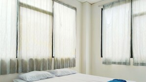 Une chambre avec un lit simple, du linge de lit blanc, deux fenêtres avec des rideaux légers et une serviette bleue sur le lit.