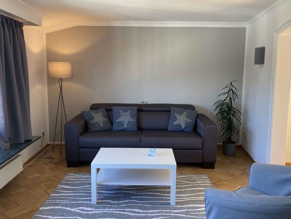 A living room with a dark grey sofa, a white coffee table, a blue armchair, a floor lamp, and a potted plant.