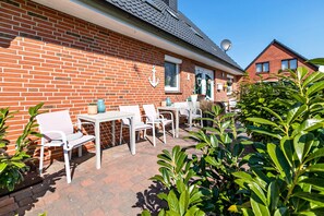 Een gebouw van baksteen met een patio, uitgerust met witte stoelen en tafels, omringd door groen.