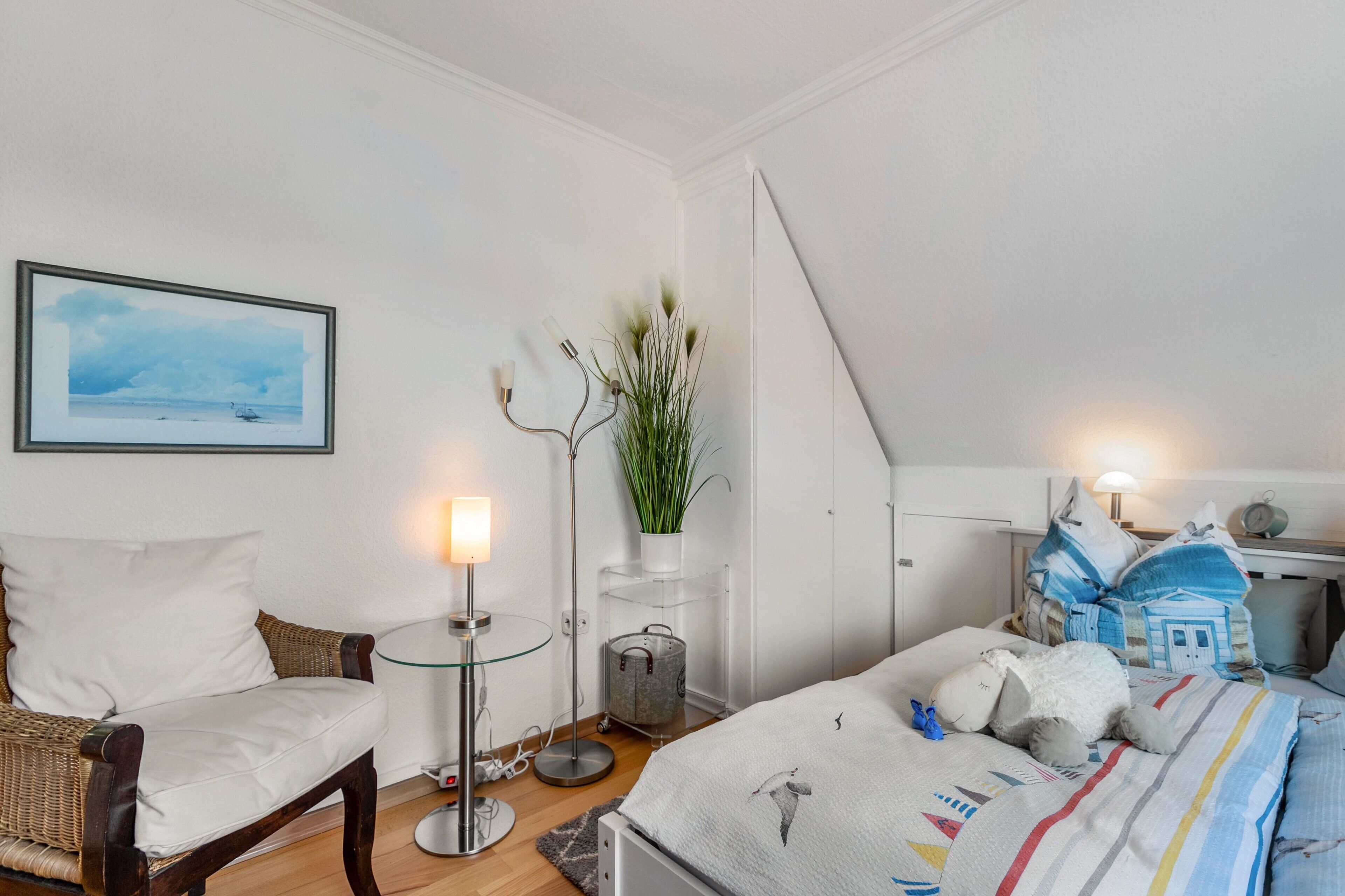 A bedroom with a bed, a chair, a bedside table, a lamp, a plant, and a framed picture on the wall.
