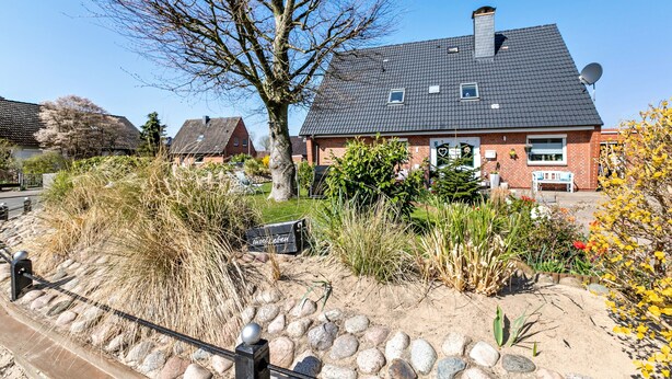 Una casa residenziale con facciata in mattoni, tetto scuro e un giardino con diverse piante e un albero.