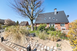Une maison d’habitation avec une façade en briques, un toit foncé et un jardin agrémenté de diverses plantes et d’un arbre.