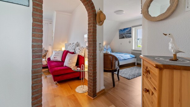 A cozy room with a red sofa, a wooden dresser, and a mirror. - Apartment 'Wattlöwe Tönning' with Private Terrace, Private Garden and Wi-Fi (Tönning)