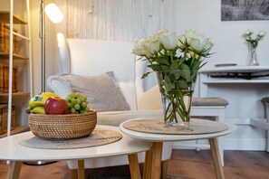 Una habitación con un sofá blanco, una mesa redonda con una cesta de mimbre llena de fruta y un jarrón de rosas blancas.