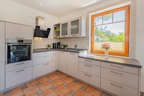 Une cuisine moderne avec des meubles blancs, un four encastré, une fenêtre avec des stores et une plante en pot posée sur le rebord de la fenêtre.