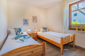 A bedroom with two single beds, a wooden nightstand with a lamp, a window with a view of greenery, and two framed pictures on the wall.