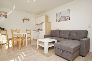 Un salon moderne avec un canapé gris, des chaises en bois, une table basse blanche et un coin cuisine équipé d’un lave-vaisselle et de meubles de rangement en bois.