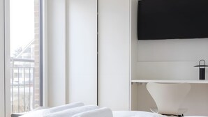 A hotel room with a bed, a flat-screen TV mounted on the wall, a white chair, and a window with a view of buildings. - Apartment 'Steuerbord' with Balcony and Wi-Fi (Sankt Peter-Ording)