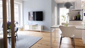 A modern living room with a flat-screen TV, a dining area with a white table and chairs, and a kitchen area with a hanging light fixture. - Apartment 'Steuerbord' with Balcony and Wi-Fi (Sankt Peter-Ording)