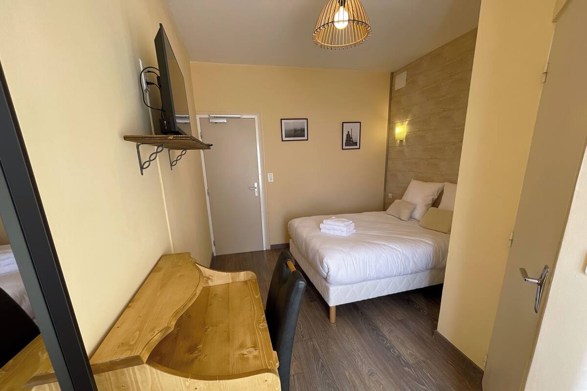 Quarto de hotel compacto com uma cama, um banco de madeira, uma mesa de trabalho e uma televisão fixada na parede.