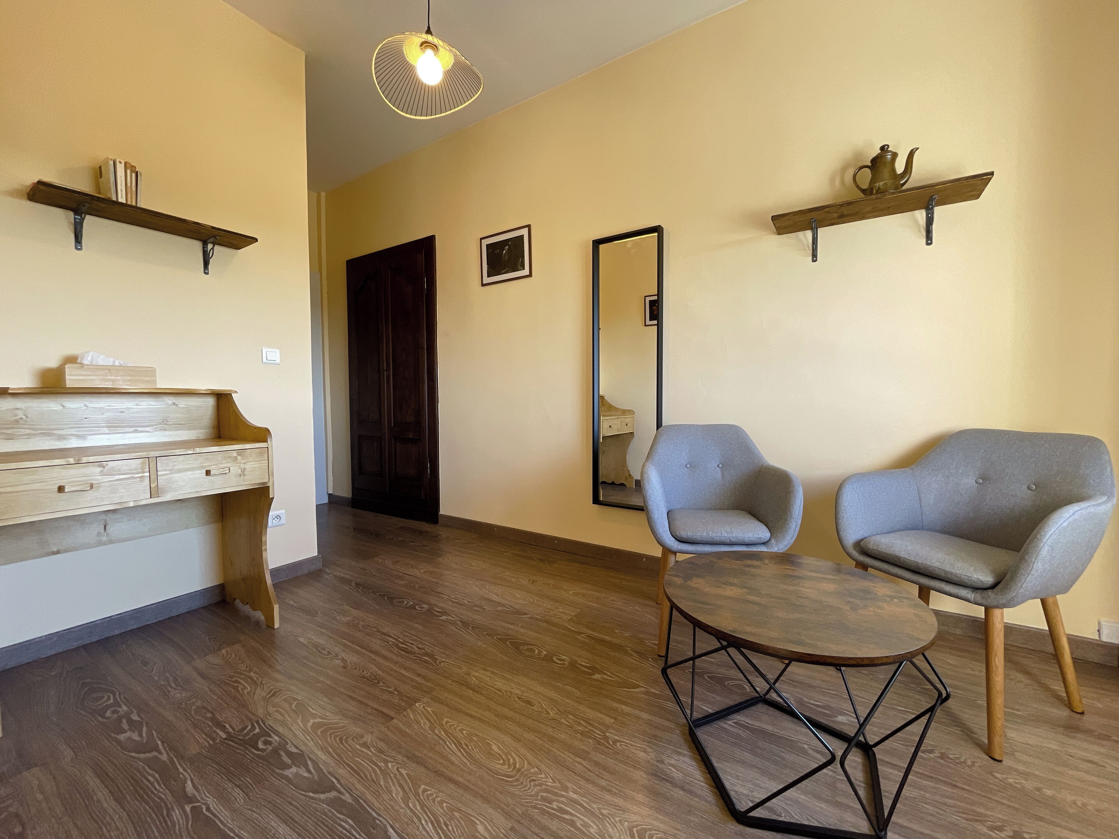 A modern living room with a wooden floor, a round table, two grey armchairs, a wooden console table with drawers, a large mirror, and two wall-mounted shelves with decorative items.