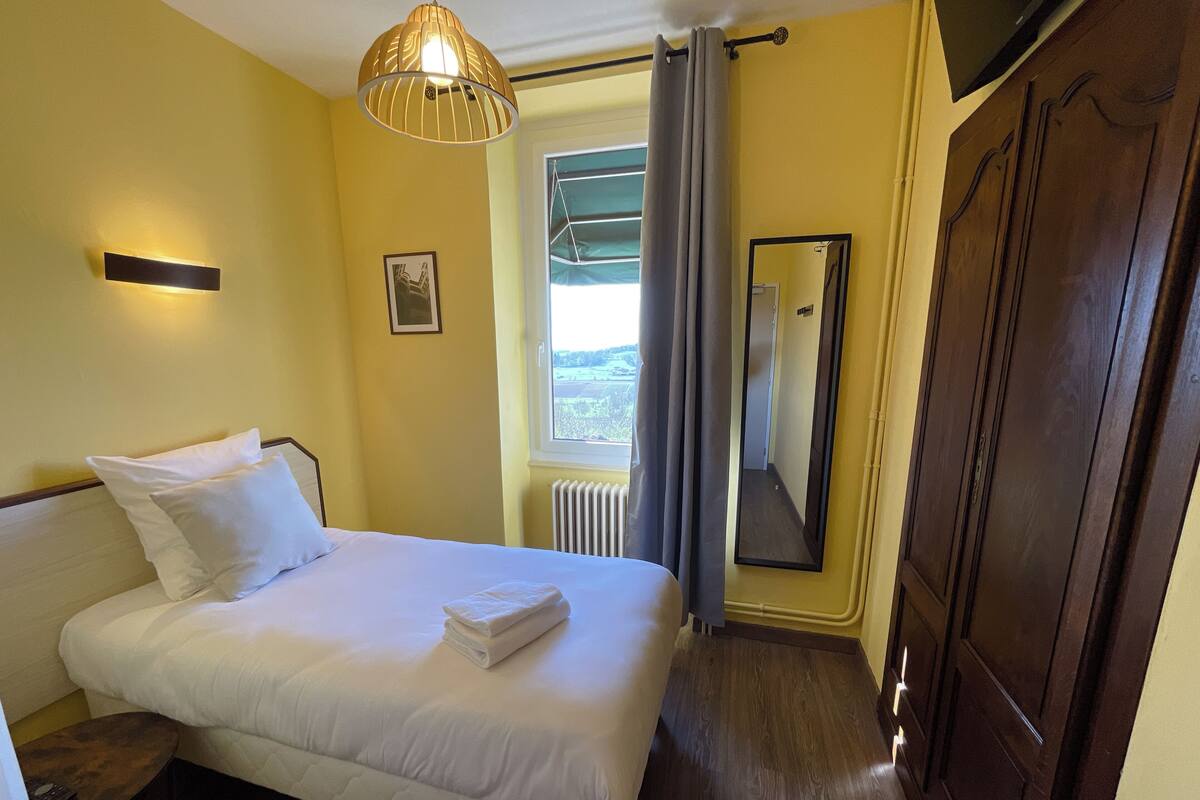 Quarto de hotel com cama, mesa de cabeceira, espelho, janela com cortinas e radiador.