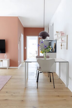 Un salon moderne avec un téléviseur, des meubles blancs, une table à manger et un vase rempli de fleurs.