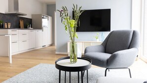 A modern living room with a grey armchair, a round black coffee table with a vase of green plants, a flat-screen TV mounted on the wall, and a kitchen in the background. - Apartment 'Backbord' with Balcony and Wi-Fi (Sankt Peter-Ording)