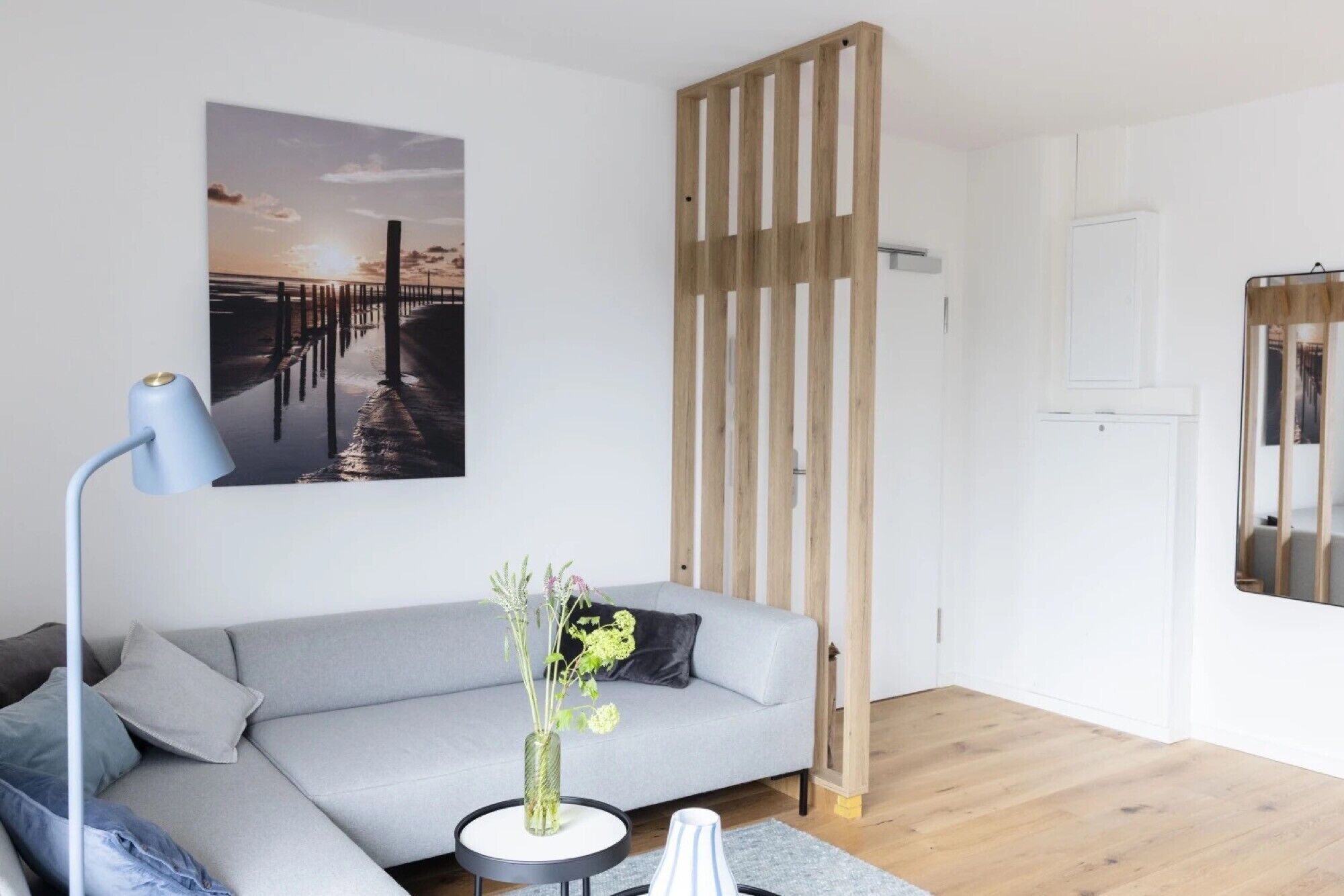 Ein modernes Wohnzimmer mit einem grauen Sofa, einem runden Beistelltisch, einer Stehlampe, einer Holzvertierung und einem gerahmten Bild an der Wand.