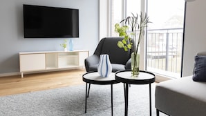 A modern living room with a flat-screen TV, a grey sofa, two round tables, and a large window with a view. - Apartment 'Backbord' with Balcony and Wi-Fi (Sankt Peter-Ording)