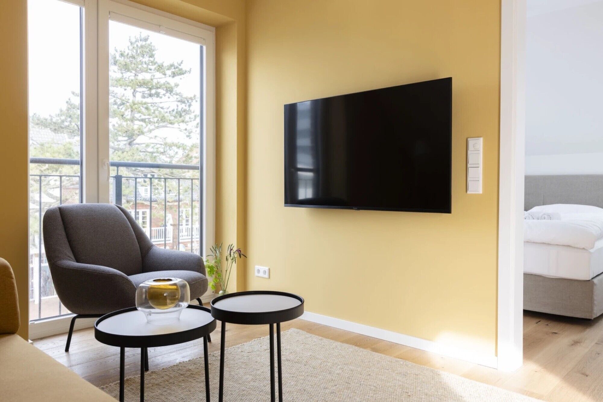 Habitación de hotel moderna con un televisor grande, un sillón cómodo, una mesita con un florero y una cama con ropa de cama blanca.