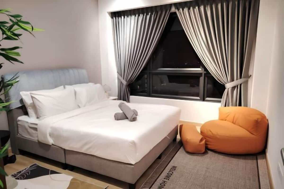 A neatly made bed with white linens, a grey headboard, and a grey pillow. There is a small orange bean bag chair and a grey rug with 'WELCOME HOME' text.