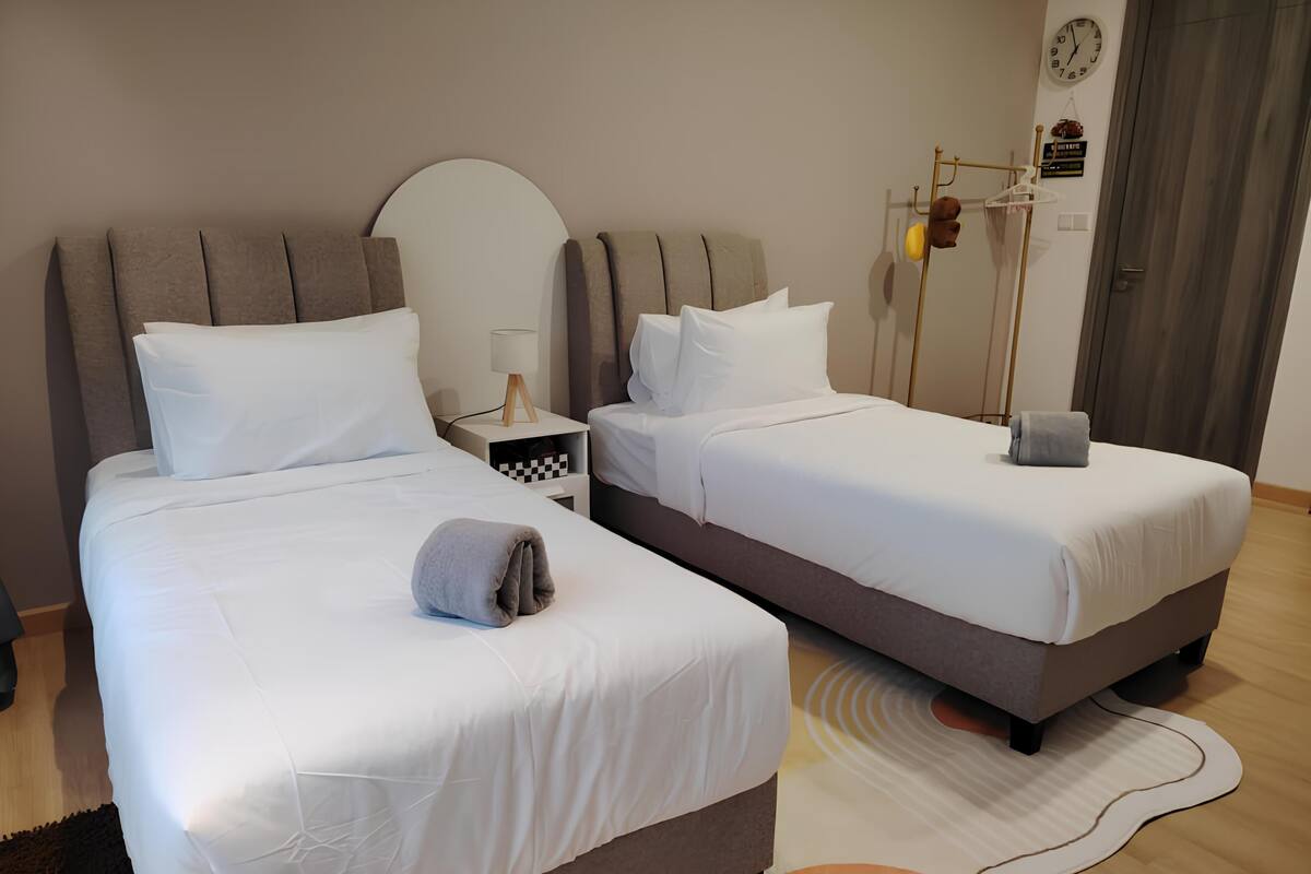 A hotel room with two beds, a bedside table with a lamp, a wall clock, and a coat rack with a teddy bear.