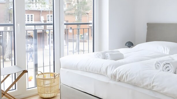 Une chambre moderne avec un grand lit, un plancher en bois et une vue sur l’extérieur, accessible par une porte-fenêtre donnant sur un balcon.