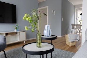 Una sala de estar moderna con una mesa redonda negra, un florero de vidrio con plantas verdes, un florero azul y un televisor de pantalla plana montado en la pared.