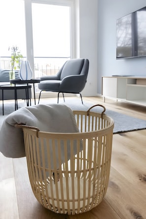 A modern living room with a wicker basket, grey armchair, and a flat-screen TV mounted on the wall.