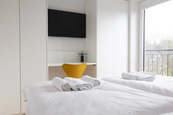 A hotel room with two beds, a desk with a chair, a mounted TV, and a balcony view.
