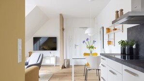 Une cuisine moderne avec des armoires blanches, un comptoir noir et un four encastré. On y trouve un coin repas avec une table et des chaises blanches, ainsi qu’un téléviseur fixé au mur.