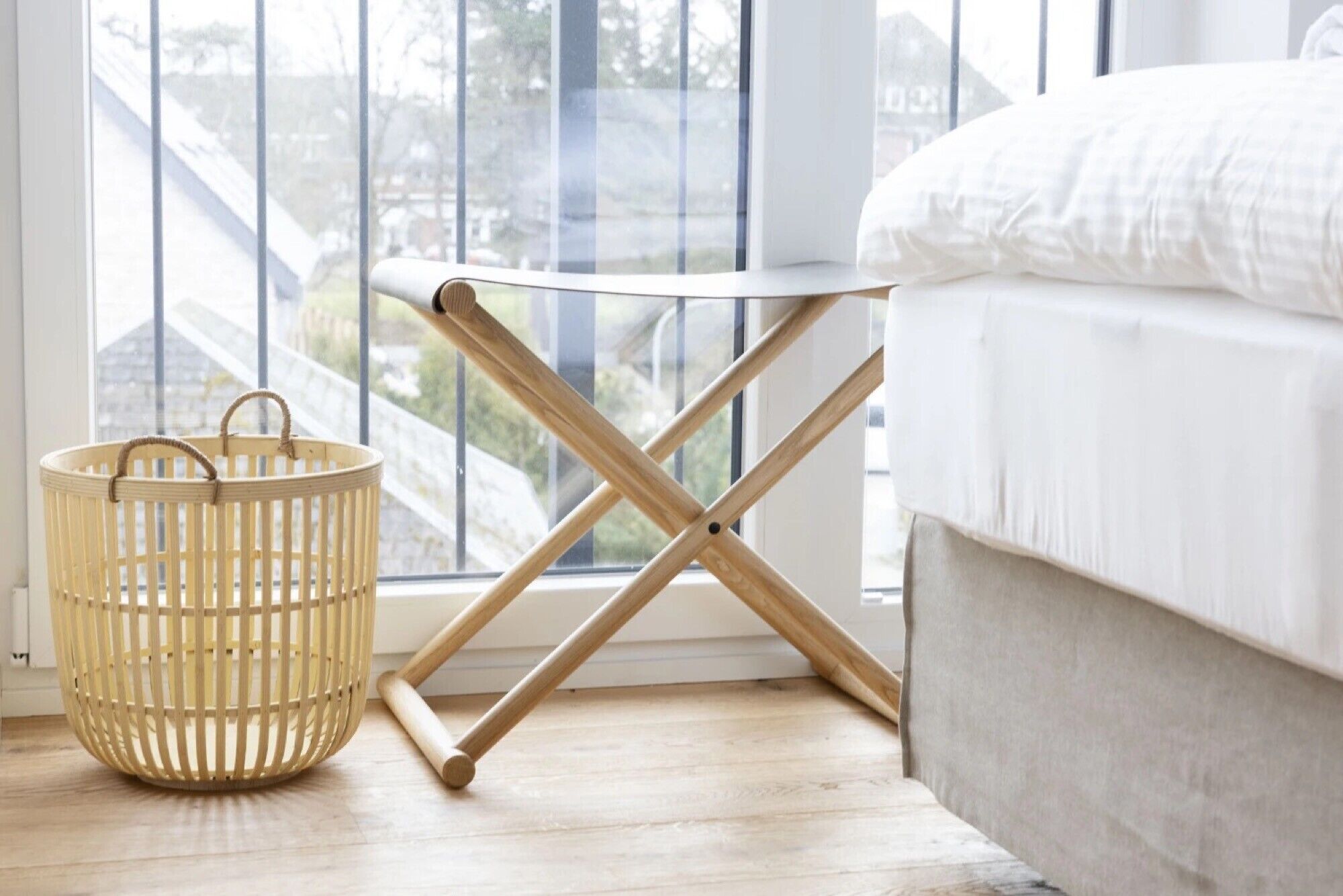 A bed with white bedding, a wooden floor, a window with a view of buildings, and a woven basket with handles.