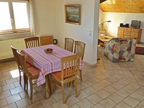 Zona pranzo con tavolo imbandito per quattro persone, sedie di legno, un termosifone, un quadro appeso al muro e una televisione su un mobile in legno.