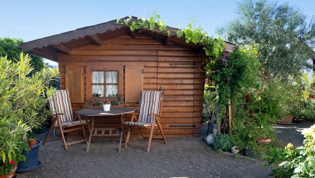 A wooden cabin with a round table and two striped chairs, surrounded by potted plants and a garden.
